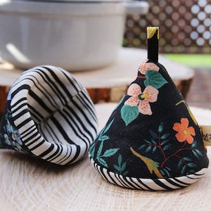 Peut inclure: Deux maniques rayées noir et blanc. L'une est dépliée pour montrer l'intérieur, qui est doublé d'un tissu floral. L'autre est fermée et présente un motif floral avec des fleurs roses, orange et vertes sur fond noir. Les maniques ont une boucle en tissu noir sur le dessus.