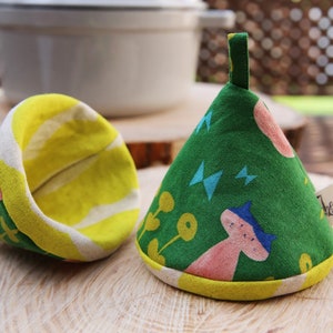 May include: Two green and yellow fabric pot holders with a whimsical pattern of pink mushrooms, blue stars, and yellow flowers. The pot holders are shaped like cones and have a loop handle.
