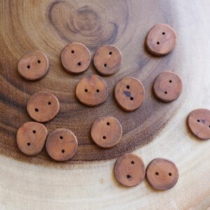 May include: A collection of thirteen circular brown wooden buttons, each featuring two small holes in the centre. The buttons exhibit natural wood grain patterns and varying shades of light to medium brown, with smooth, rounded edges throughout.