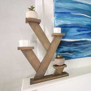 May include: A wooden plant stand with a unique V-shaped design. The stand has three shelves, each holding a small potted plant. The wood has a natural, warm tone. The stand is placed against a white wall with a blue painting in the background.