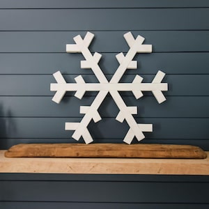 Puede incluir: Una gran decoración de copo de nieve de madera blanca. El copo de nieve está montado en una pared azul oscuro sobre una estantería de madera. El copo de nieve tiene seis brazos simétricos, cada uno con tres extensiones rectangulares.