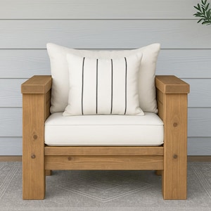 Könnte beinhalten: Ein hölzerner Outdoor-Sessel mit einem hellen Kissen und zwei Kissen. Ein Kissen hat vertikale schwarze Streifen. Der Stuhl ist aus hellbraunem Holz gefertigt und steht auf einem grauen Teppich. Der Hintergrund ist eine hellblaue Wand.