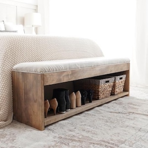 以下が含まれることがあります： 明るい色の布張りの座面が付いた木製のベンチ。ベンチの下部には靴と2つの織りバスケットが置かれた棚があります。木材は自然な仕上がりで、ベッドの足元に配置されています。