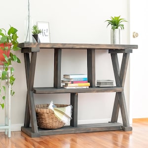 May include: A dark stained wooden console table with two shelves and a cross-beam design. The top shelf holds a small potted plant, a framed print with the text "hi!", and a glass vase. The lower shelf displays books and a woven basket.