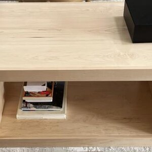 Puede incluir: Una mesa de centro de madera de color claro con una superficie rectangular y un estante de almacenamiento abierto debajo. Una caja rectangular negra s&oacute;lida se encuentra sobre la superficie superior, mientras que una pila de libros descansa en el estante.
