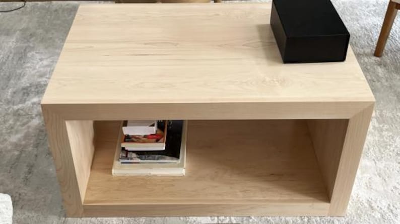 Puede incluir: Una mesa de centro de madera de color claro con una superficie rectangular y un estante de almacenamiento abierto debajo. Una caja rectangular negra s&oacute;lida se encuentra sobre la superficie superior, mientras que una pila de libros descansa en el estante.