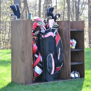 Puede incluir: Unidad de almacenamiento de palos de golf de madera con tinte oscuro. La unidad tiene un compartimento para bolsas de golf y estantes para pelotas y zapatos de golf. Las bolsas de golf son negras y rojas.