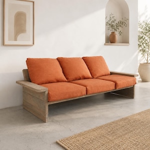 Pode incluir: Um sofá de três lugares com uma estrutura de madeira castanho-clara com acabamento natural e três almofadas grossas de cor laranja vibrante. O sofá tem apoios de braços planos e largos e um design baixo, proporcionando um assento estruturado.