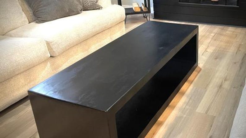 Puede incluir: Una mesa de centro rectangular con un acabado negro s&oacute;lido y una superficie superior lisa. La mesa cuenta con un compartimento de almacenamiento abierto que atraviesa el centro, creando un espacio hueco entre la parte superior y la base.