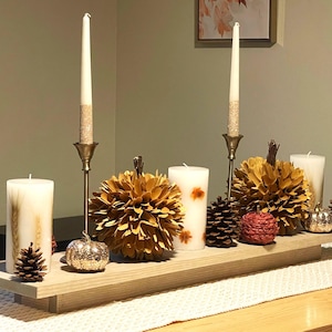 Puede incluir: Centro de mesa otoñal con dos velas altas en candelabros dorados, dos calabazas decorativas y piñas. El arreglo se exhibe sobre una bandeja de madera, creando un ambiente cálido y acogedor.