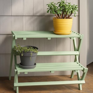 Op de afbeelding: Een tweekleurige, lichtgroene houten plantenstandaard met twee potplanten. De bovenste plank heeft een gele pot met een groene plant en de onderste plank heeft een grijze pot met een groene plant.