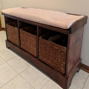 May include: A dark brown wooden storage bench with three woven baskets and a cushioned top. The cushion is beige with red stripes. The bench is designed for storage and seating.