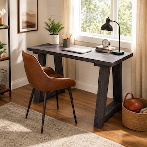 Peut inclure: Un bureau en bois gris anthracite foncé avec un plateau rectangulaire et des pieds inclinés, accompagné d'une chaise en similicuir couleur cognac avec des pieds en métal noir. Le bureau porte un ordinateur portable argenté et une lampe.