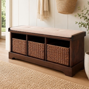 May include: A dark brown wooden storage bench featuring three open cubbies, each containing a woven wicker basket. The top of the bench is fitted with a rectangular, tufted seat cushion upholstered in a light beige fabric with thin brown stripes.