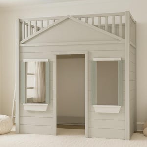 Puede incluir: Una cama con forma de casa de madera gris claro con un marco en forma de casa, dos aberturas en forma de ventana y un pequeño balcón. Un banderín rosa con la palabra "MAGIC" cuelga de la pared y un oso de peluche beige está sentado cerca. Un otomano blanco está en primer plano.