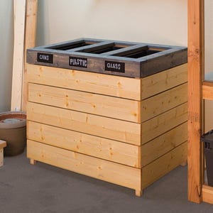 May include: A wooden recycling bin with three compartments labelled "CANS", "PLASTIC", and "GLASS". The bin is made from light-coloured wood with a dark grey top. A practical and stylish solution for waste management.