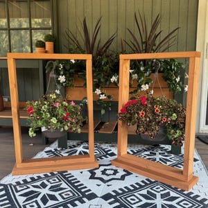 May include: Two wooden hanging plant stands with rectangular frames. Each stand holds a hanging basket filled with colorful flowers. The stands are on a black and white patterned rug, with a green background and a bench visible in the background.