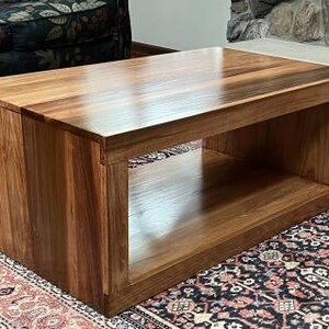 Puede incluir: Una mesa de centro rectangular construida en madera marr&oacute;n pulida con un patr&oacute;n de veta visible. La mesa cuenta con una superficie superior s&oacute;lida y un gran compartimento de almacenamiento abierto debajo, sostenido por dos paneles laterales verticales gruesos.