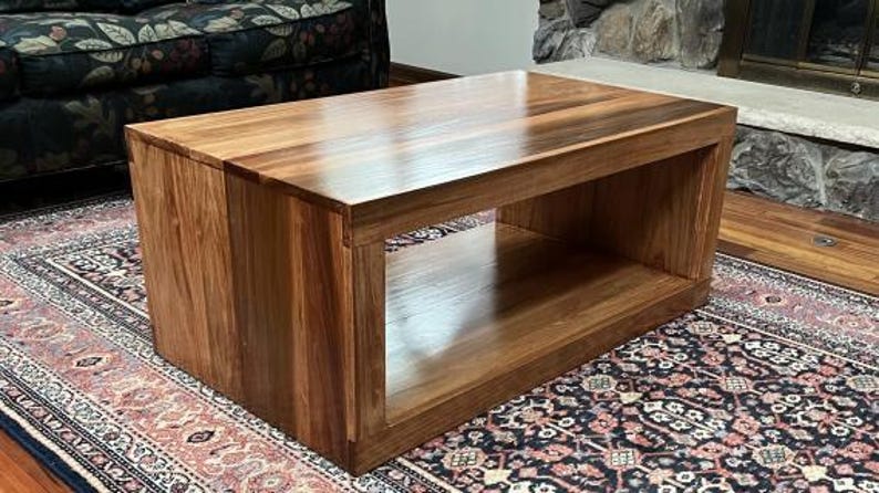 Puede incluir: Una mesa de centro rectangular construida en madera marr&oacute;n pulida con un patr&oacute;n de veta visible. La mesa cuenta con una superficie superior s&oacute;lida y un gran compartimento de almacenamiento abierto debajo, sostenido por dos paneles laterales verticales gruesos.