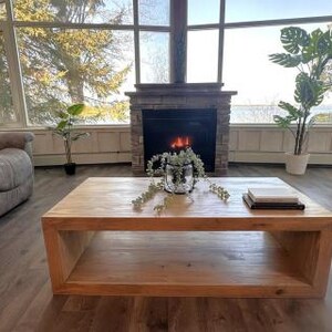 Puede incluir: Una mesa de centro rectangular de madera marr&oacute;n claro con un dise&ntilde;o de estante central hueco. La mesa presenta un acabado liso de veta de madera natural y se asienta sobre un suelo de aspecto de madera oscura en una habitaci&oacute;n soleada.