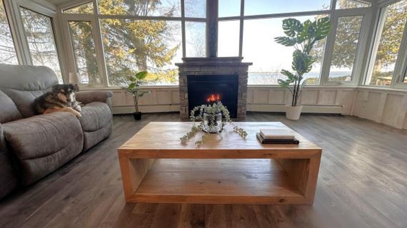 Puede incluir: Una mesa de centro rectangular de madera marr&oacute;n claro con un dise&ntilde;o de estante central hueco. La mesa presenta un acabado liso de veta de madera natural y se asienta sobre un suelo de aspecto de madera oscura en una habitaci&oacute;n soleada.