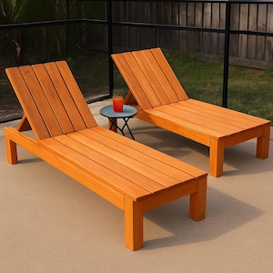 May include: Two wooden chaise lounge chairs with adjustable backs. The chairs are made of light brown wood and are positioned on a concrete patio. A small blue table with a drink sits between the chairs. The background includes a fence and greenery.