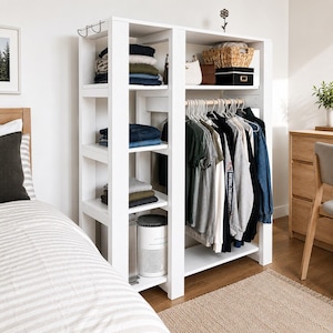 Op de afbeelding: Een witte open kledingkast met een verticaal schapgedeelte aan de linkerkant en een kledingroede aan de rechterkant. De planken bevatten opgevouwen kleding, een gevlochten mand en een kleine luchtreiniger. Aan hangers hangen diverse shirts.