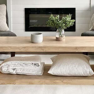 Puede incluir: Una mesa de centro rectangular de madera clara con un estante inferior. La superficie superior sostiene un peque&ntilde;o cuenco gris y un jarr&oacute;n de cristal con follaje verde. El estante inferior contiene un textil blanco doblado y un coj&iacute;n blanco.