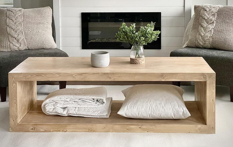 Puede incluir: Una mesa de centro rectangular de madera clara con un estante inferior. La superficie superior sostiene un peque&ntilde;o cuenco gris y un jarr&oacute;n de cristal con follaje verde. El estante inferior contiene un textil blanco doblado y un coj&iacute;n blanco.