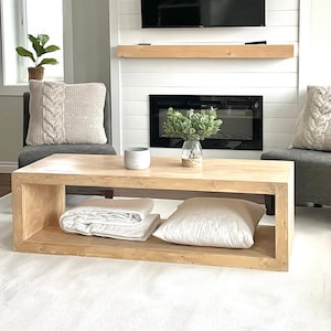 May include: A light-coloured wooden coffee table with a lower shelf holding a folded blanket and a pillow. The table sits on a white rug in front of a fireplace. Two grey chairs with cable-knit pillows are on either side.