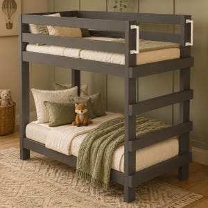 May include: A dark gray wooden bunk bed featuring a sturdy frame with horizontal slats, white safety handles on the top bunk, and two mattresses dressed in cream-colored bedding with olive green throw pillows and a textured green knit blanket.
