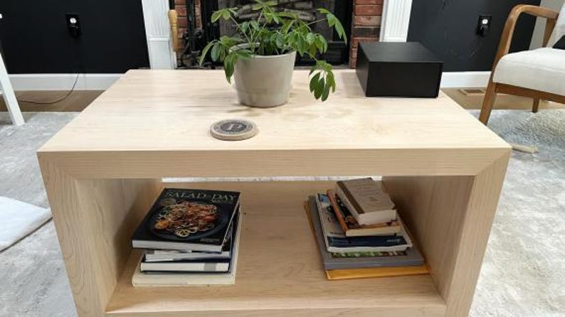 Puede incluir: Una mesa de centro de madera clara con tablero cuadrado y estante inferior abierto. El tablero sostiene una planta verde en maceta, un posavasos circular con la letra P y una caja negra. El estante inferior contiene pilas de libros y revistas.