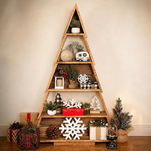 May include: A triangular wooden shelving unit shaped like a Christmas tree with five horizontal tiers. The shelves are decorated with various items including white snowflake cutouts, small artificial pine trees, wrapped gifts, and pinecones.