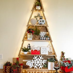May include: Wooden Christmas tree shelf decorated with festive items. The shelves hold miniature trees, a camper, snowflake ornaments, and gifts. The tree is lit with fairy lights and topped with a star.