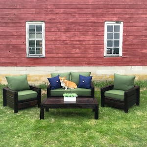 Puede incluir: Juego de muebles de exterior de madera oscura, que incluye un sofá de dos plazas, dos sillones y una mesa de centro. Los cojines son verdes y hay dos cojines azules marino. Un perro corgi está sentado en el sofá. El fondo es un granero rojo.
