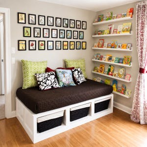Peut inclure: Un banc de rangement blanc avec trois paniers tressés noirs, surmonté d'un matelas matelassé marron foncé, de coussins verts à motifs et d'un coussin décoratif central. Au-dessus, une galerie murale affiche des cadres sur l'alphabet.