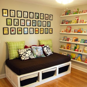 May include: A children's reading corner with a brown daybed, cushions, and storage baskets. Framed alphabet prints adorn the wall. White shelves are filled with colourful children's books, creating a comfortable space for reading and learning.