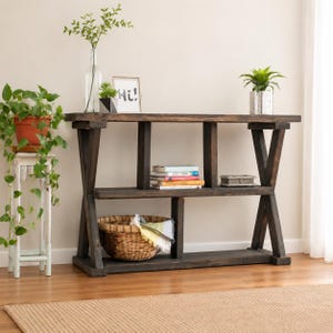 May include: A dark brown wooden console table with an X-shaped frame, featuring two horizontal shelves. The top shelf holds a stack of books and a small box, while the bottom shelf contains a woven basket with a cloth inside.