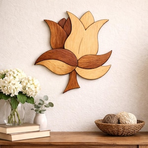 May include: A wooden flower wall art piece with a layered design in shades of brown and tan. The flower is mounted on a white wall. A vase of white flowers, books, and a basket with decorative balls are on a wooden table.