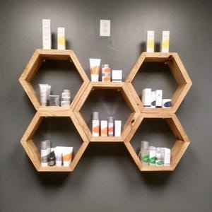 Könnte beinhalten: Holzregale in sechseckiger Form an einer grauen Wand, die verschiedene Hautpflegeprodukte präsentieren. Die Regale sind aus hellem Holz gefertigt und in einem Wabenmuster angeordnet. Die Produkte umfassen Tuben, Flaschen und Schachteln mit weißer, orangefarbener und gelber Verpackung.