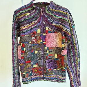 Peut inclure: Veste patchwork colorée avec fermeture éclair intégrale. La veste présente une variété de textures et de couleurs, notamment le violet, le rose, le jaune et le vert. Les manches sont longues et la veste est suspendue à un cintre blanc.