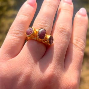 May include: A gold ring featuring three oval-shaped, deep red gemstones. The ring has a thick band and a textured gold setting, creating a bold and elegant design. The gemstones are set close together, enhancing the ring's visual impact.