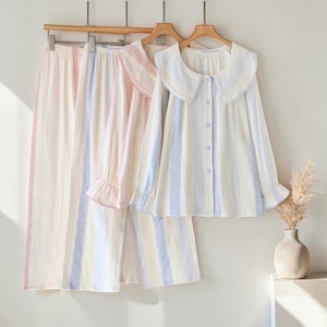 Peut inclure: Trois ensembles de pyjamas aux couleurs pastel suspendus à des cintres en bois. Les ensembles comprennent des chemises à manches longues avec un col arrondi et un devant boutonné, et des pantalons assortis. Les pyjamas ont des rayures verticales roses, bleues et blanches.