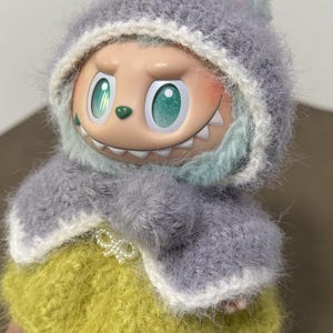 Puede incluir: Una mu&ntilde;eca caprichosa con una chaqueta con capucha gris difusa con orejas de conejo y una falda amarilla. La mu&ntilde;eca tiene ojos grandes y expresivos y una sonrisa traviesa. El atuendo est&aacute; hecho a mano, con un enfoque en la textura y los detalles.