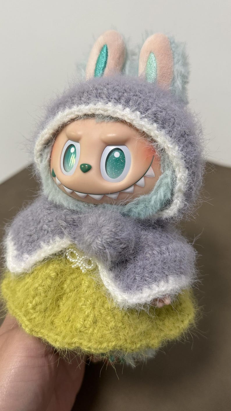 Puede incluir: Una mu&ntilde;eca caprichosa con una chaqueta con capucha gris difusa con orejas de conejo y una falda amarilla. La mu&ntilde;eca tiene ojos grandes y expresivos y una sonrisa traviesa. El atuendo est&aacute; hecho a mano, con un enfoque en la textura y los detalles.