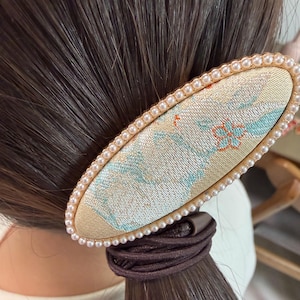 May include: Oval hair clip with a pearl border. The clip features a beige fabric with a floral pattern in blue and orange. The clip is attached to a brown hair tie and is set against a background of brown hair.