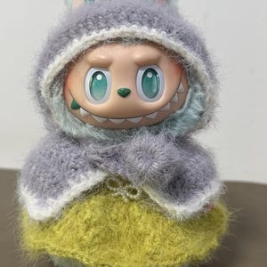 Puede incluir: Una figura caprichosa de una criatura con ojos grandes y una sonrisa dentuda, vestida con un atuendo de punto. Presenta una capa con capucha gris y una falda amarilla. La figura tiene orejas de conejo y una expresi&oacute;n juguetona.