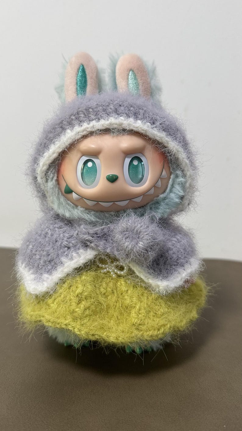 Puede incluir: Una figura caprichosa de una criatura con ojos grandes y una sonrisa dentuda, vestida con un atuendo de punto. Presenta una capa con capucha gris y una falda amarilla. La figura tiene orejas de conejo y una expresi&oacute;n juguetona.