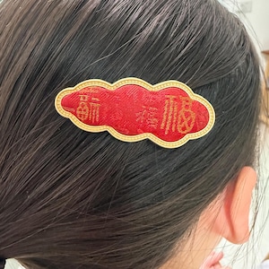 Op de afbeelding: Rode, wolk-vormige haarclip met gouden rand en Chinese karakters. De clip is gemaakt van een textuurstof en is bevestigd aan donker haar. Het ontwerp is waarschijnlijk voor een speciale gelegenheid.