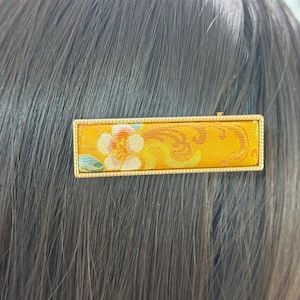 May include: A rectangular hair clip with a gold frame and a yellow fabric insert. The fabric features a floral design with orange, white, and green accents. The clip is attached to brown hair.